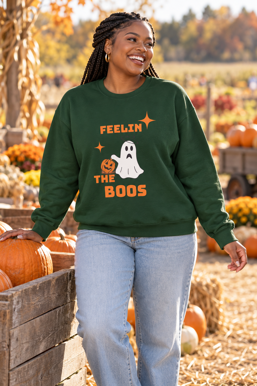 A woman wearing a dark green crewneck sweatshirt with a Halloween graphic that reads 'FEELIN' THE BOOS' and shows a ghost and pumpkin. She is standing in an outdoor pumpkin patch.