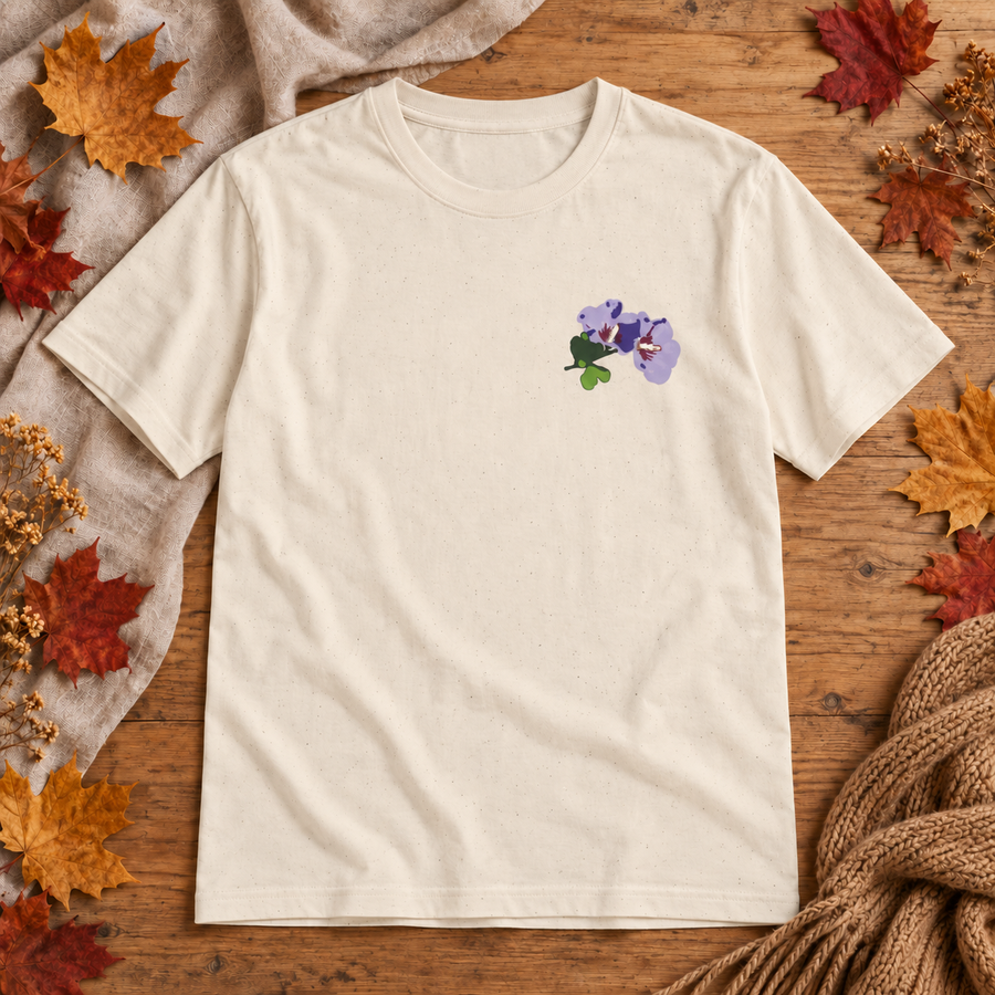 A beige short-sleeve crewneck t-shirt with a small purple and green flower graphic on the left chest, laid flat on a rustic wooden surface surrounded by autumn leaves, a beige blanket, and a knitted sweater.