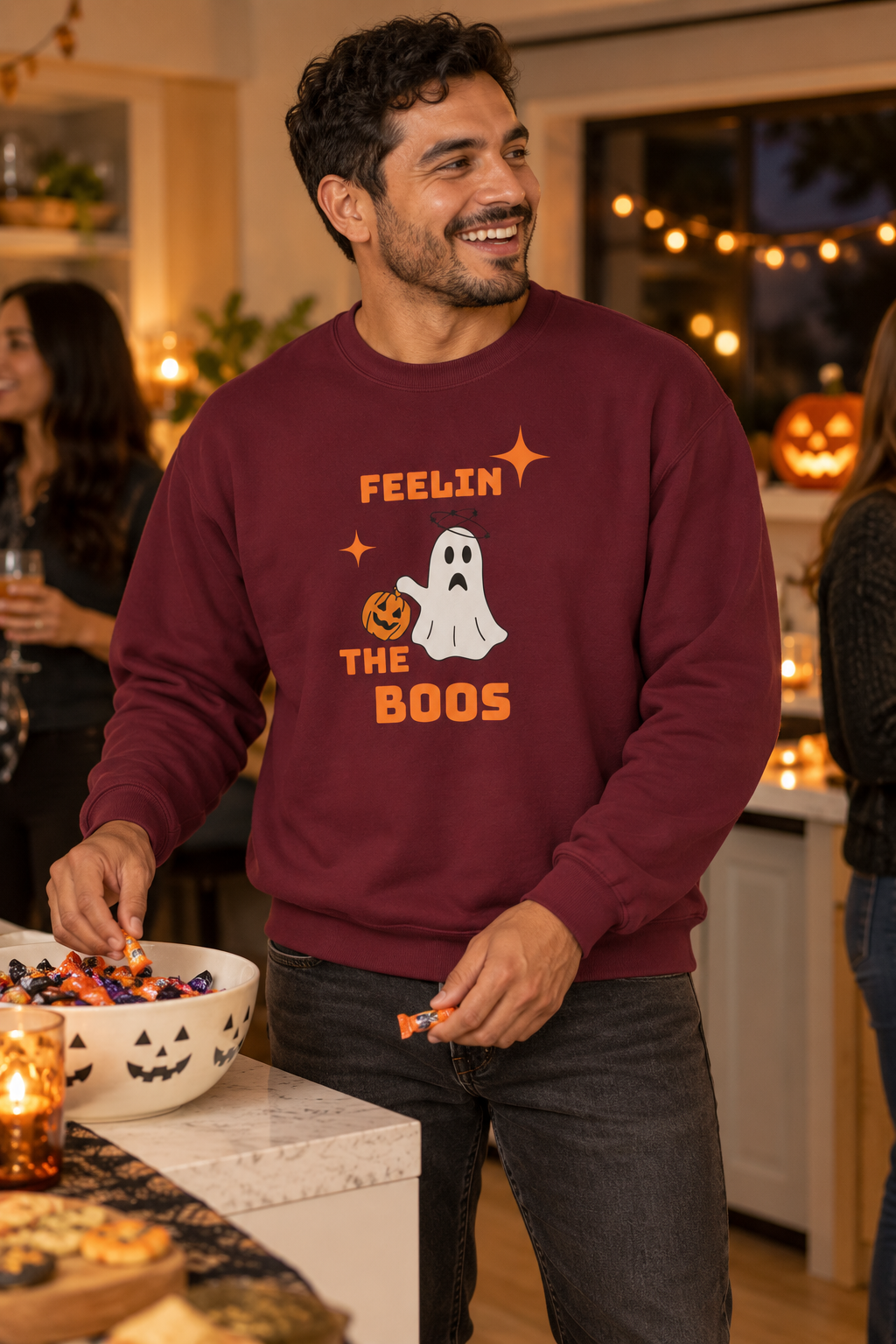 A man wearing a maroon crewneck sweatshirt with a Halloween graphic that reads 'FEELIN THE BOOS' with a ghost and pumpkin, standing in a warmly lit holiday party kitchen.