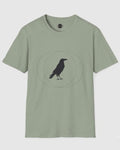 A sage green short-sleeve crewneck t-shirt featuring a black crow silhouette standing inside a geometric circle graphic on the chest, displayed against a light gray background.