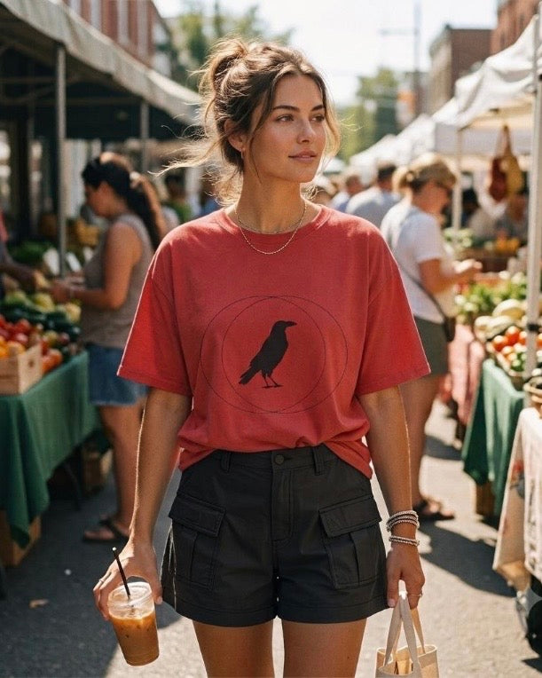 A woman walking through an outdoor farmers market wearing a coral-red graphic t-shirt with a crow design and black cargo shorts, holding an iced coffee.
