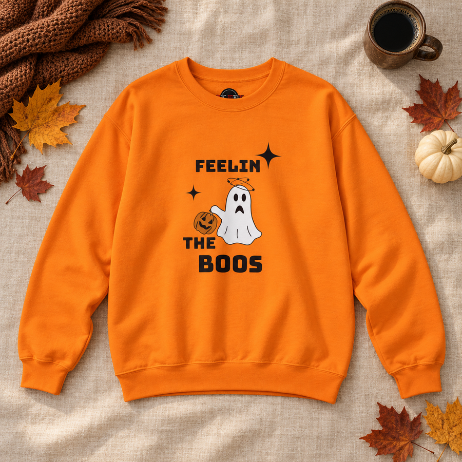 An orange crewneck sweatshirt with a Halloween graphic featuring a white ghost, a jack-o'-lantern, and the text 'FEELIN THE BOOS' in black. The sweatshirt is displayed in a flat-lay arrangement on a beige linen background, surrounded by autumn leaves, a brown knit scarf, a small pumpkin, and a coffee cup.