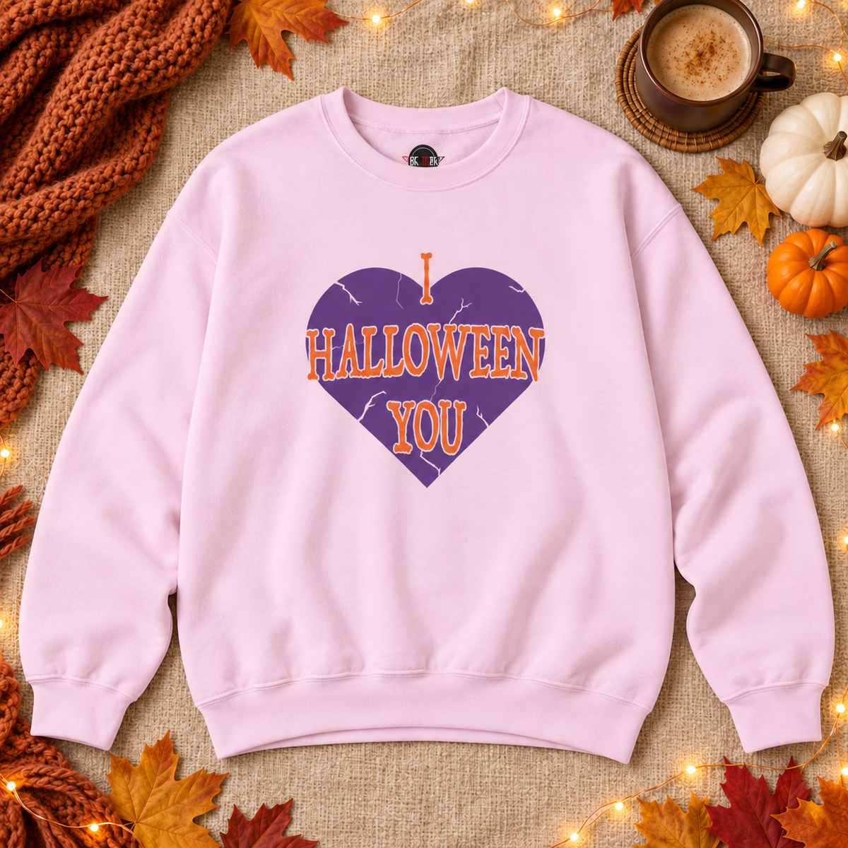 A light pink crewneck sweatshirt laid flat on a burlap surface, featuring a purple heart graphic with orange text that reads 'I HALLOWEEN YOU.' The scene is styled with autumn leaves, a knitted orange scarf, a cup of coffee, and small pumpkins.