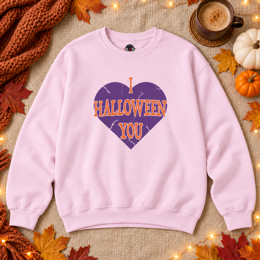 A light pink crewneck sweatshirt laid flat on a burlap surface, featuring a purple heart graphic with orange text that reads 'I HALLOWEEN YOU.' The scene is styled with autumn leaves, a knitted orange scarf, a cup of coffee, and small pumpkins.