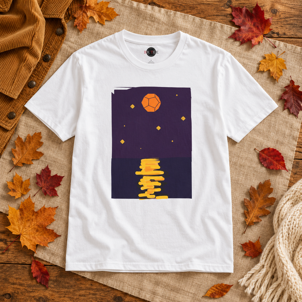 A white short-sleeve crewneck t-shirt with a graphic print of a geometric orange moon and yellow stylized text on a dark purple background, styled flat-lay on a burlap cloth with autumn leaves and a brown corduroy jacket.